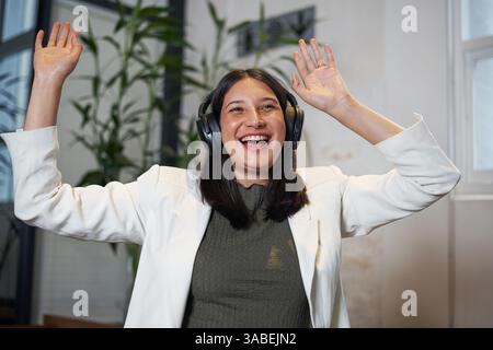 Lächelnde Frau, die Kopfhörer trägt und die Hände hebt, in einem hellen Innenbereich Stockfoto
