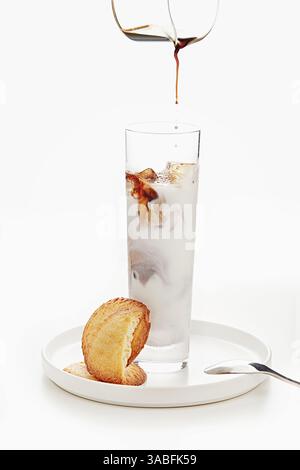 Nahaufnahme eines Kaffees, der in ein hohes Glas Eismilch gegossen wird, um einen Eislatt und Kekse auf einem Teller zu machen Stockfoto