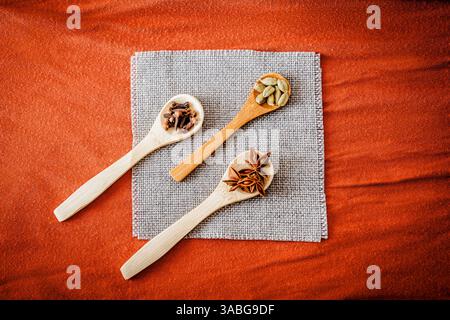 Drei Holzlöffel mit saisonalen Gewürzen auf Sackleinen-Serviette und warmen orangefarbenen Stoffen. Rustikale kulinarische Flachlage, ideal für Herbst-Food-Blogs und Bio-Co Stockfoto