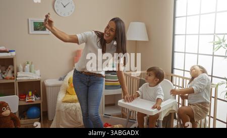 Frau nimmt Selfie mit Sohn und Tochter in einem gemütlichen Schlafzimmer, in dem Liebe und Familienbande in der Nähe einer Wiege präsentiert werden, umgeben von einem verspielten und warmen Zuhause Stockfoto