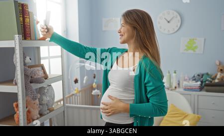 Schwangere Frau nimmt Selfie im gemütlichen Kinderzimmer mit Kinderbett, umgeben von Babyspielzeug und Essentials, um fröhliche Momente in Innenräumen festzuhalten. Stockfoto
