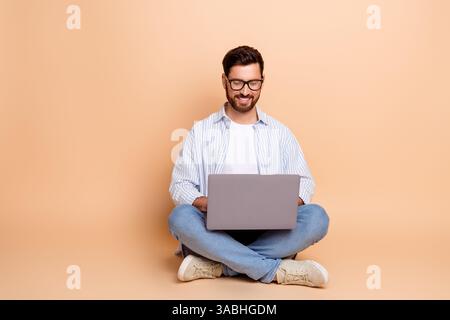 Junger Mann, der einen Laptop benutzt und im Kreuz auf beigefarbenem Hintergrund sitzt und ein lässiges blau gestreiftes Hemd mit Brille trägt Stockfoto