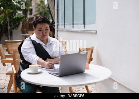 Eine junge asiatische Frau in Übergröße sitzt vor einem Café und konzentriert sich auf ihren Laptop. Umgeben von Grün arbeitet sie an der Arbeit und genießt die Plädoyer Stockfoto