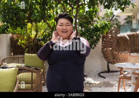 Eine junge asiatische Frau in Übergröße sitzt selbstbewusst vor einem Café und trägt ein stilvolles Outfit. Sie legt ihr Kinn auf die Hand, während sie zur Seite blickt. Stockfoto