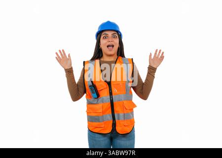 Überraschte junge Bauarbeiterin mit orangefarbener Sicherheitsweste und blauem Helm hebt ihre Hände mit aufgerissenem Mund und drückt aus Stockfoto