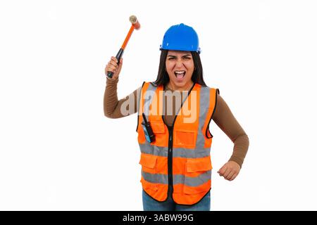 Wütende lateinische Bauarbeiterin, die eine orange Weste und einen blauen Helm trägt, einen Hammer hält und schreien, zeigt Frustration und Stärke gegenüber Stockfoto