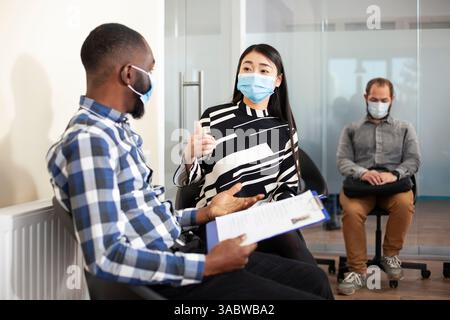 Arbeitssuchende mit cvs, die sich in der Lobby unterhalten, darunter asiatische Frau, die mit schwarzem Mann spricht, die beide maskiert sind und auf Interviews warten. Verschiedene Kandidaten halten die Zwischenablage und sprechen vor dem Einstellungsgespräch. Stockfoto