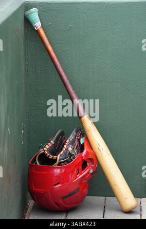 Baseballausrüstung einschließlich Schläger, Helm und Handschuh in der Ecke eines Dugouts während eines Baseballspiels. Stockfoto
