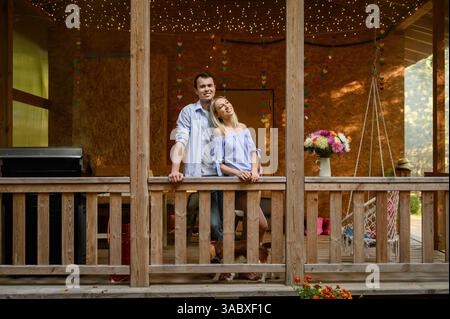 Liebevolles glückliches Familienpaar steht auf der Veranda des Hauses aneinander, lächelt glücklich, Hund rennt in der Nähe. Konzept von Romantik, Unterstützung, Träumen, Re Stockfoto
