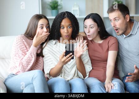 Erstaunliche Gruppe von Freunden, die überraschende Inhalte auf einem Telefon auf einer Couch zu Hause überprüfen Stockfoto