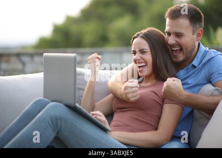 Aufregendes Paar, das Inhalte von Notebooks überprüft und auf einer Terrasse zu Hause feiert Stockfoto