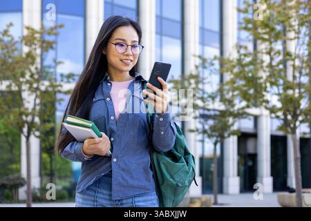 Eine Asiatin mit Brille überprüft ihr Handy, während sie auf dem Campus läuft, Bücher hält und einen Rucksack trägt. Stockfoto