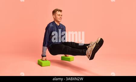 Junger Mann mit erhöhtem L-Sit auf Schaumstoffblöcken, mit blauem Sportoberteil und schwarzer Hose, zeigt Stärke und Kernkraft vor rosa Hintergrund. Stockfoto