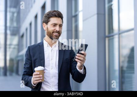 Erfolgreicher Geschäftsmann, der mit dem Telefon in der Hand durch die Stadt spaziert und glücklich lächelt. Mann im Business-Anzug, der in sozialen Medien surft und Textnachrichten schreibt, mit einer Tasse Heißgetränk in der Hand. Stockfoto