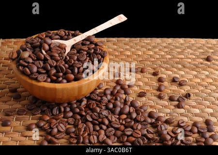 Braune Kaffeebohnen, die aus einer Holzschale mit einem Löffel ausgeschüttet werden, liegen auf einer gewebten Matte unter weicher, warmer Beleuchtung, die die natürlichen Strukturen unterstreicht Stockfoto