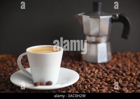 Espresso in einer weißen Keramiktasse, umgeben von gerösteten Kaffeebohnen und einer traditionellen Espressomaschine mit Herd für ein authentisches Kaffeeerlebnis Stockfoto