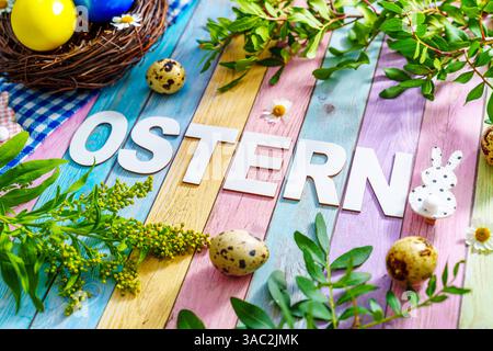 Bayern, Deutschland - 2. April 2025: Der Schriftzug OSTERN auf farbigem Holzuntergrund, umgeben von Osterdekorationen, frischem Grün und einem Osternest mit bunten Eiern *** der Schriftzug OSTERN auf farbigem Holzuntergrund, umgeben von Osterdekoration, frischem Grün und einem Osternest mit bunten Eiern Stockfoto