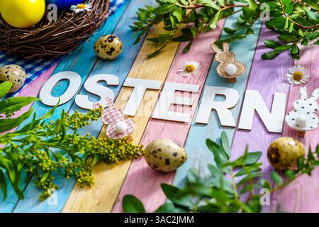 Bayern, Deutschland - 2. April 2025: Der Schriftzug OSTERN auf farbigem Holzuntergrund, umgeben von Osterdekorationen, frischem Grün und einem Osternest mit bunten Eiern *** der Schriftzug OSTERN auf farbigem Holzuntergrund, umgeben von Osterdekoration, frischem Grün und einem Osternest mit bunten Eiern Stockfoto