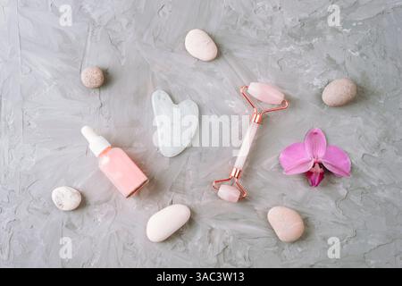 Flache Lagerung von Hautpflegewerkzeugen einschließlich Gua Sha, Gesichtsrolle, Serumtropfflasche und rosa Orchidee auf grauem Hintergrund. Draufsicht. Stockfoto