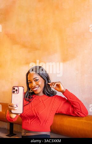 Eine junge Frau, die mit ihrem Smartphone in einem Café ein Selfie macht, lächelt und ihr Haar berührt Stockfoto