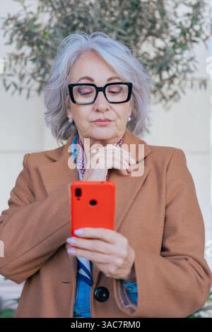 Stilvolle ältere kaukasische Frau mit Smartphone und Denken im Freien Stockfoto