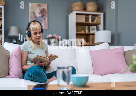 Junge Frau in lässigen Klamotten auf der Couch, hört Musik mit kabellosen Kopfhörern und liest ein Buch. Zeit zu lesen, Buchliebhaber, weiblich, genießt W Stockfoto