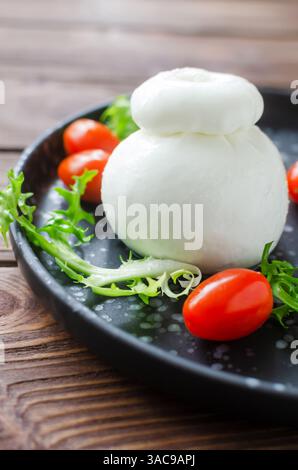 Buratta auf einem schwarzen Teller mit Kirschtomaten und Kräutern auf einem Holztisch. Traditionelles italienisches Essenskonzept. Vertikale Ausrichtung. Selektiver Fokus. Stockfoto
