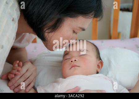 Nahaufnahme einer Mutter, die ihr friedlich schlafendes Baby sanft auf die Stirn küsst, während sie die Hand des Kindes in einer hölzernen Krippe hält Stockfoto