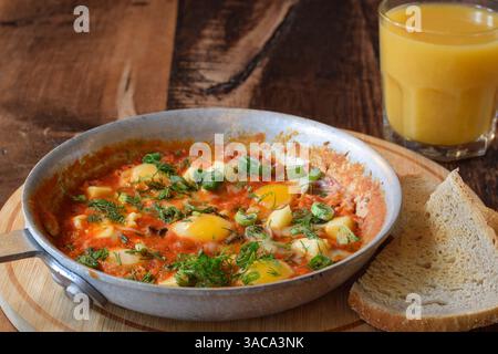 Frühstück Shakshuka garniert mit frischem Dill und grünen Zwiebeln in der Pfanne auf Holztisch mit einem Glas Orangensaft und einer Seite Brot Toast Stockfoto