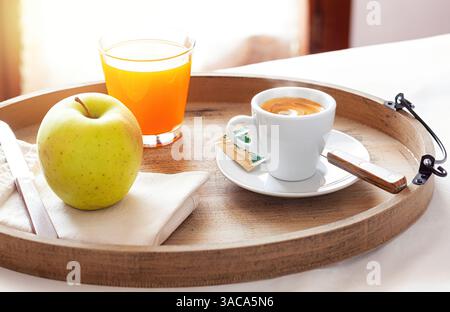 Holztablett mit frischem Frühstück mit Kaffee, Fruchtsaft und knusprigem Apfel, serviert in einem gemütlichen Hotelzimmer, das sich im warmen Sonnenlicht sonnt und für ein perfektes Erlebnis am Morgen sorgt Stockfoto