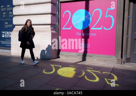 Am Morgen nach der Ankündigung von europäischen und weltweiten Handelszöllen durch US-Präsident Trump, die eine Abgabe von 10 % für importierte Waren aus Großbritannien beinhalteten, läuft ein Käufer am 3. April 2025 in London, England, an einer Plakatwand für das Jahr 2025 auf Piccadilly im Londoner West End vorbei. Präsident Trumps Ankündigung einer langen Liste von Zöllen gilt als die größte Umwälzung des Welthandels seit einem Jahrhundert. Mit der Vorhersage, dass es einen globalen Handelskrieg auslösen wird. Stockfoto