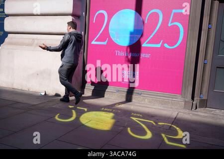 Am Morgen nach der Ankündigung von europäischen und weltweiten Handelszöllen durch US-Präsident Trump, die eine Abgabe von 10 % für importierte Waren aus Großbritannien beinhalteten, läuft ein Käufer am 3. April 2025 in London, England, an einer Plakatwand für das Jahr 2025 auf Piccadilly im Londoner West End vorbei. Präsident Trumps Ankündigung einer langen Liste von Zöllen gilt als die größte Umwälzung des Welthandels seit einem Jahrhundert. Mit der Vorhersage, dass es einen globalen Handelskrieg auslösen wird. Stockfoto