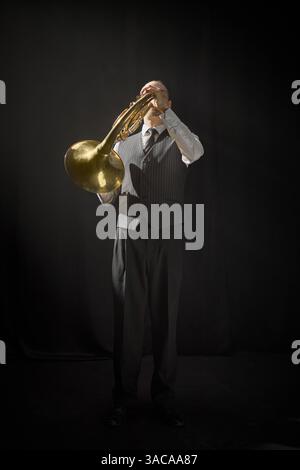 Vertikale Aufnahme eines männlichen Musikers in formeller Weste und eleganter Krawatte, der Horn spielt, der vor schwarzem Hintergrund steht, Kopierraum Stockfoto