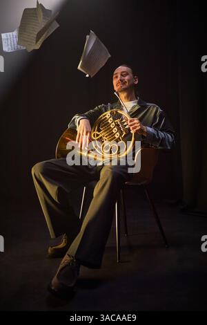 Vertikale Aufnahme eines professionellen männlichen Musikers, der im Studio mit Horn sitzt, während Notenblätter in der Luft vor schwarzem Hintergrund schweben, Inspirationskonzept für Komponisten Stockfoto