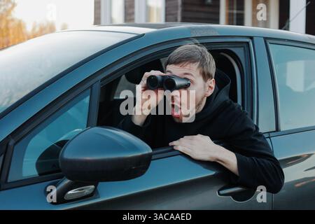 Junger, schockierter Stalker, der durch das Fernglas im Auto sitzt. Mann spioniert oder ermittelt. Stockfoto