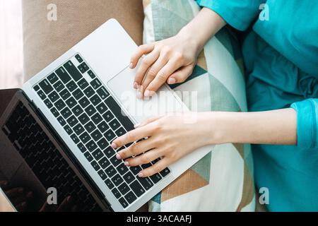Nahaufnahme von Frauenhänden, die auf einem Laptop tippen, während sie mit einem gemusterten Kissen auf einer Couch sitzen. Weiche Produktivität und neue Arbeitsatmosphäre, Fernarbeit Stockfoto