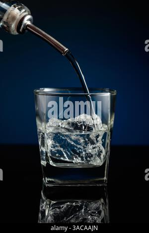 Kristallklarer Wodka oder Gin wird über Eis in ein Glas gegossen. Wodka mit Eis auf schwarzem reflektierendem Hintergrund. Stockfoto