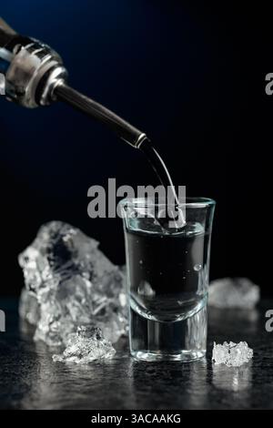 Kristallklarer Wodka oder Gin wird in ein Glas gegossen. Transparentes alkoholisches Getränk mit Eisstücken auf schwarzem Hintergrund. Stockfoto