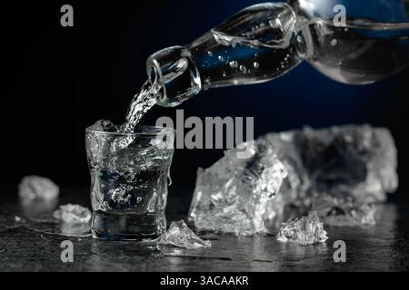 Kristallklarer Wodka oder Gin wird in ein Glas gegossen. Transparentes alkoholisches Getränk mit Eisstücken auf schwarzem Hintergrund. Stockfoto