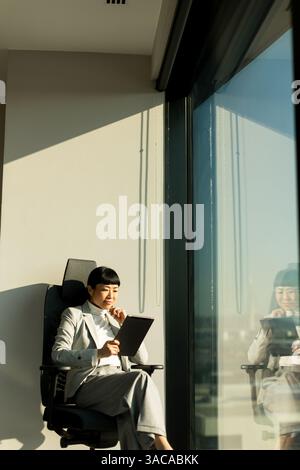 In einem sonnigen Bürobereich sitzt eine japanische Geschäftsfrau in formeller Kleidung bequem in einem modernen Stuhl. Sie konzentriert sich auf ihr Tablet und reflektiert ein Stockfoto