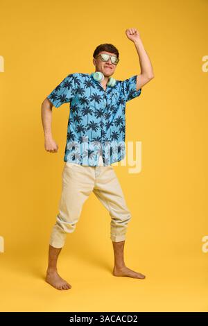 Ganzkörperfoto eines ausdrucksstarken jungen Mannes, der barfuß mit Kopfhörern am Hals tanzt und den Moment des Urlaubs genießt. Stockfoto