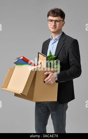 Junger Geschäftsmann mit ernstem Ausdruck, der Karton mit Büchern und Pflanzen hält. Konzept des unerwarteten Arbeitsplatzwechsels Stockfoto