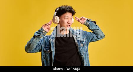 Junger Mann in Jeansjacke und Kopfhörern, der mit ausdrucksstarkem Gesicht auf gelbem Hintergrund zum Beat tanzt. Stockfoto