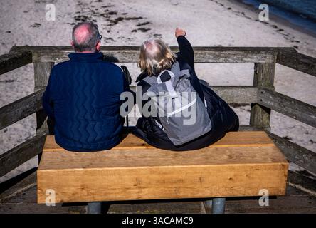 Boltenhagen, Deutschland. April 2025. Ein pensioniertes Paar sitzt auf einer Holzbank in der Sonne am Pier mit Blick auf die Ostseeküste. Quelle: Jens Büttner/dpa/Alamy Live News Stockfoto