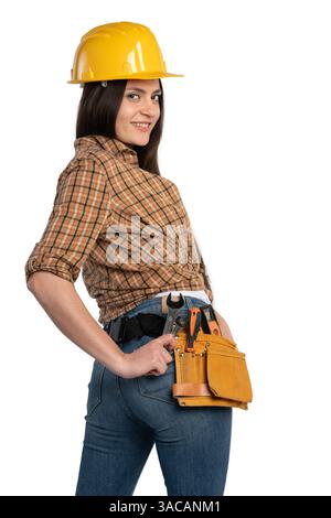 Porträt einer lächelnden Bauarbeiterin mit gelbem Helm, kariertem Hemd, Jeans und Werkzeuggürtel, die selbstbewusst isoliert auf weiß posiert. Stockfoto