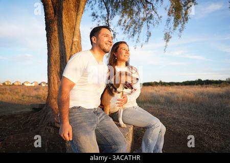 Ein fröhliches Paar, das bei Sonnenuntergang mit seinem Hund unter einem Baum sitzt und einen Moment der Liebe und Freundschaft festnimmt. Stockfoto