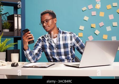 Ein junger Freelancer sitzt an seinem Schreibtisch und schaut sich sein Mobilgerät an. Ein afroamerikanischer Berater hält sein Smartphone und liest Textnachrichten, während er eine Pause von seinen Aufgaben einlegt. Stockfoto