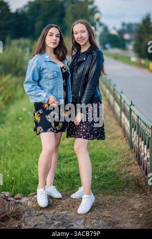 Zwei glückliche junge Frauen, die Blumenkleider und weiße Turnschuhe tragen, genießen ihre gemeinsame Zeit in einem Park Stockfoto