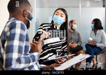 Arbeitslose, die ihren lebenslauf halten und ein Gespräch im Büro des Unternehmens führen. Asiatische Frau, die mit schwarzem Mann im Lobbybereich spricht, beide tragen Gesichtsmasken und warten darauf, interviewt zu werden. Stockfoto