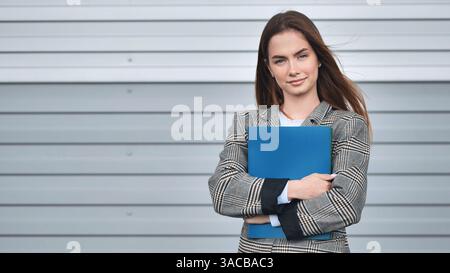 Professionelle Geschäftsfrau lächelnd, packender blauer Ordner in der Nähe eines eleganten grauen Wandhintergrunds Stockfoto